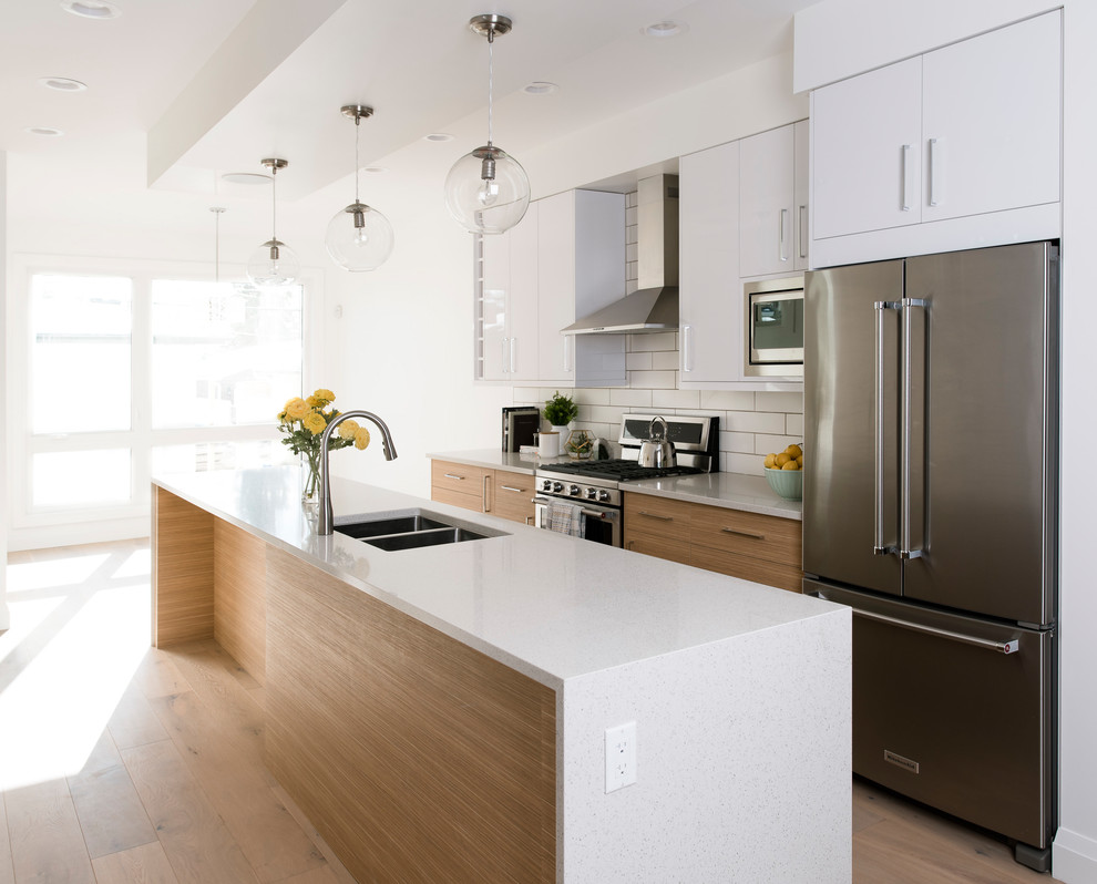 This is an example of a medium sized contemporary single-wall kitchen/diner in Edmonton with a double-bowl sink, flat-panel cabinets, white cabinets, engineered stone countertops, white splashback, metro tiled splashback, stainless steel appliances, light hardwood flooring, an island and beige floors.