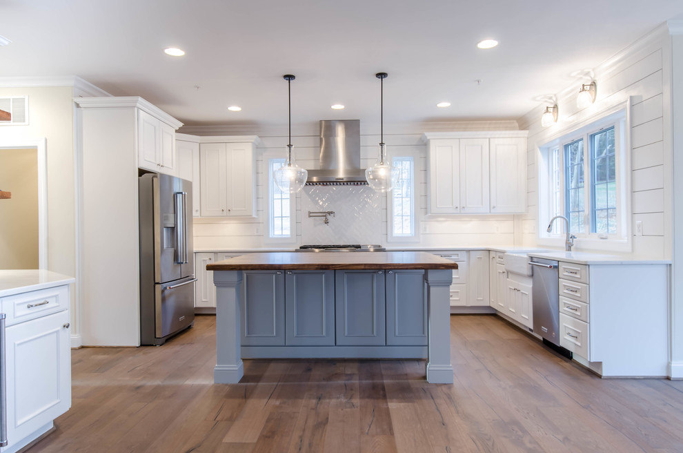 Photo of a medium sized classic u-shaped kitchen/diner in Baltimore with a belfast sink, shaker cabinets, white cabinets, engineered stone countertops, white splashback, ceramic splashback, stainless steel appliances, light hardwood flooring and an island.