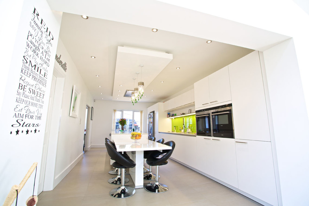 Large contemporary galley open plan kitchen in Hampshire with an integrated sink, flat-panel cabinets, white cabinets, composite countertops, green splashback, glass sheet splashback, stainless steel appliances, porcelain flooring and an island.