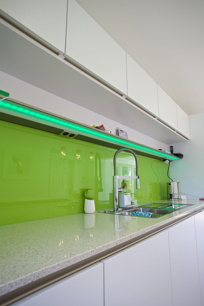 Medium sized contemporary galley kitchen/diner in Hampshire with an integrated sink, flat-panel cabinets, white cabinets, composite countertops, green splashback, glass sheet splashback, stainless steel appliances, porcelain flooring and an island.