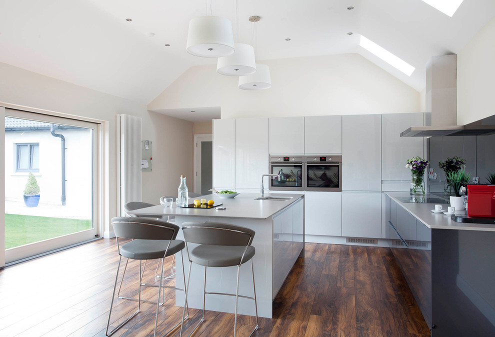 Medium sized contemporary l-shaped open plan kitchen in Other with an integrated sink, flat-panel cabinets, grey cabinets, quartz worktops, grey splashback, mirror splashback, stainless steel appliances, medium hardwood flooring and an island.