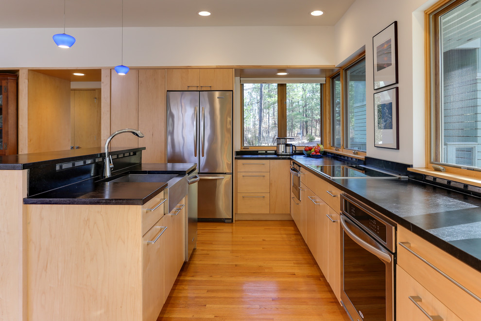 Design ideas for a medium sized contemporary l-shaped open plan kitchen in Raleigh with a belfast sink, flat-panel cabinets, light wood cabinets, soapstone worktops, stainless steel appliances, medium hardwood flooring, an island, black worktops and brown floors.