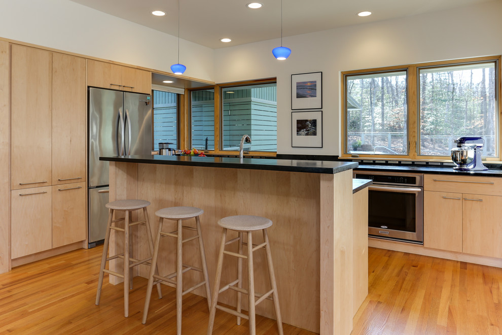 This is an example of a medium sized modern l-shaped open plan kitchen in Raleigh with a belfast sink, flat-panel cabinets, light wood cabinets, soapstone worktops, stainless steel appliances, medium hardwood flooring, an island and black worktops.