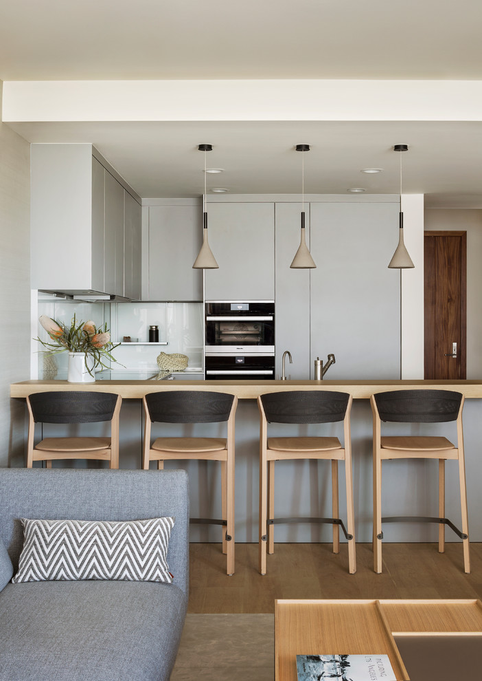 This is an example of a contemporary u-shaped open plan kitchen in Los Angeles with flat-panel cabinets, grey cabinets, wood worktops, white splashback, glass sheet splashback, light hardwood flooring and a breakfast bar.