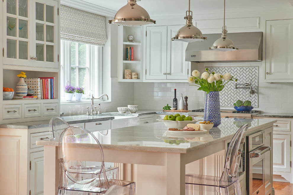 Klassisk inredning av ett vit vitt kök och matrum, med en rustik diskho, luckor med upphöjd panel, vita skåp, marmorbänkskiva, vitt stänkskydd, rostfria vitvaror, ljust trägolv, en köksö och brunt golv