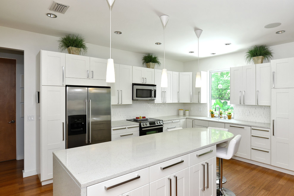 Example of a mid-sized minimalist l-shaped medium tone wood floor open concept kitchen design in Tampa with a double-bowl sink, recessed-panel cabinets, white cabinets, granite countertops, white backsplash, ceramic backsplash, stainless steel appliances and an island