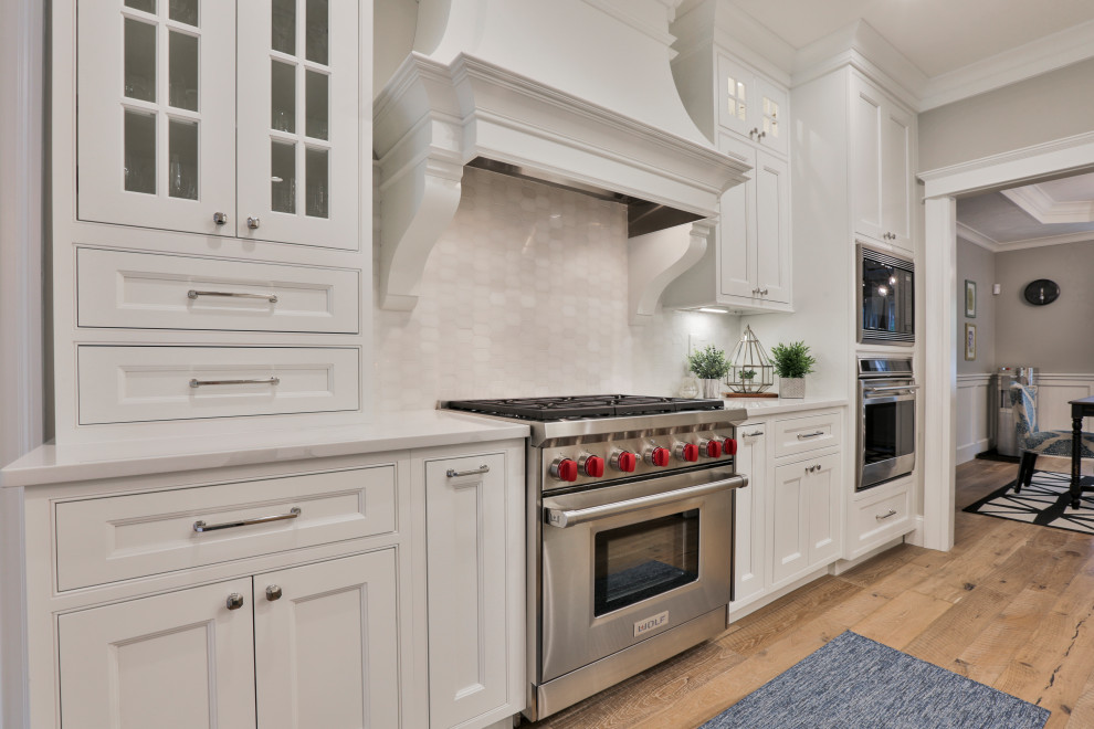 Klassisk inredning av ett stort vit vitt kök, med en rustik diskho, luckor med profilerade fronter, vita skåp, bänkskiva i kvarts, vitt stänkskydd, stänkskydd i marmor, rostfria vitvaror, ljust trägolv, en köksö och flerfärgat golv