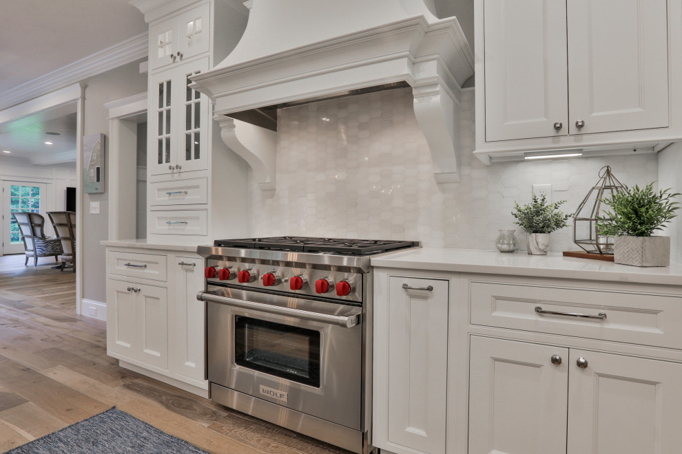 Large traditional u-shaped kitchen/diner in Boston with a belfast sink, beaded cabinets, white cabinets, engineered stone countertops, white splashback, marble splashback, stainless steel appliances, light hardwood flooring, an island, multi-coloured floors and white worktops.