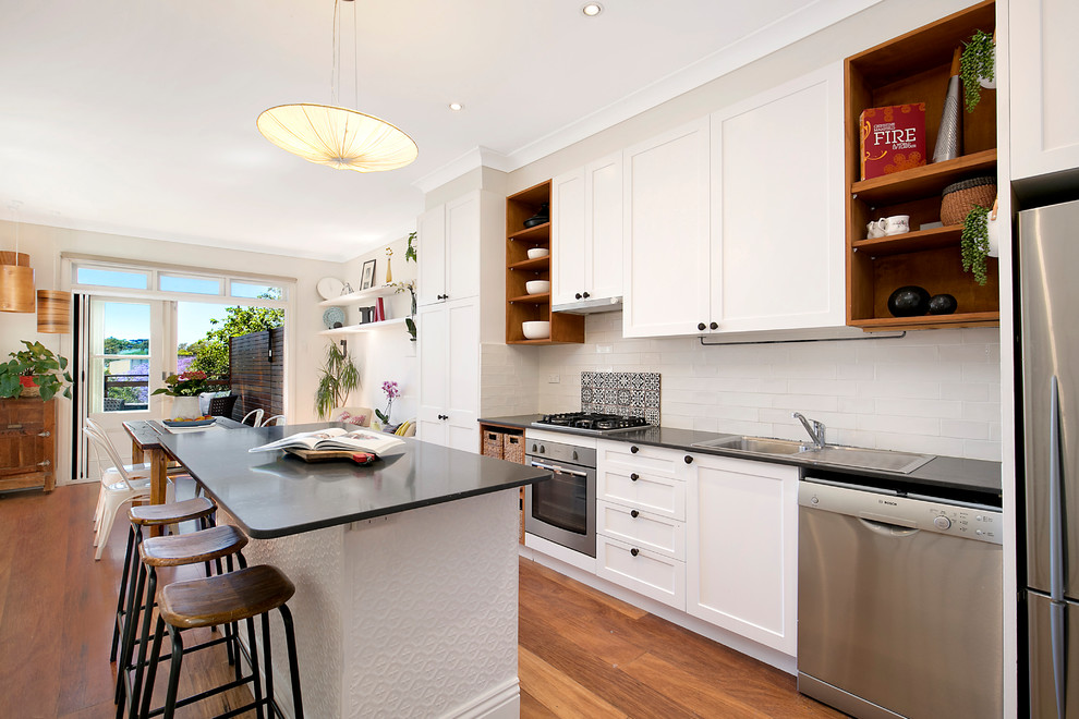 This is an example of an eclectic single-wall kitchen/diner in Sydney with a single-bowl sink, white cabinets, white splashback, metro tiled splashback, stainless steel appliances, medium hardwood flooring, an island, recessed-panel cabinets, composite countertops and black worktops.