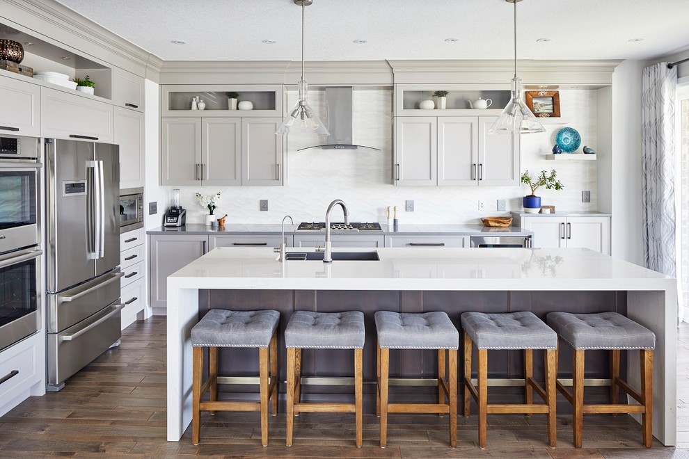 Imagen de cocina rectangular tradicional renovada grande con armarios estilo shaker, puertas de armario grises, salpicadero blanco, electrodomésticos de acero inoxidable, una isla, suelo marrón, fregadero bajoencimera, suelo de madera oscura y encimeras grises