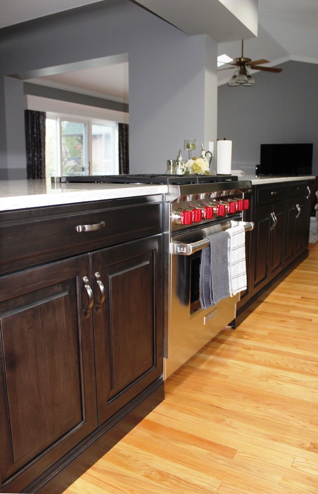This is an example of a large traditional galley open plan kitchen in Detroit with a single-bowl sink, raised-panel cabinets, white cabinets, engineered stone countertops, grey splashback, glass tiled splashback, stainless steel appliances, light hardwood flooring, an island and white worktops.
