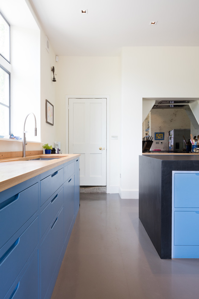 Inspiration for a l-shaped open plan kitchen in Wiltshire with flat-panel cabinets, blue cabinets, wood worktops, metallic splashback, mirror splashback, an island, grey floors, brown worktops, a submerged sink and stainless steel appliances.