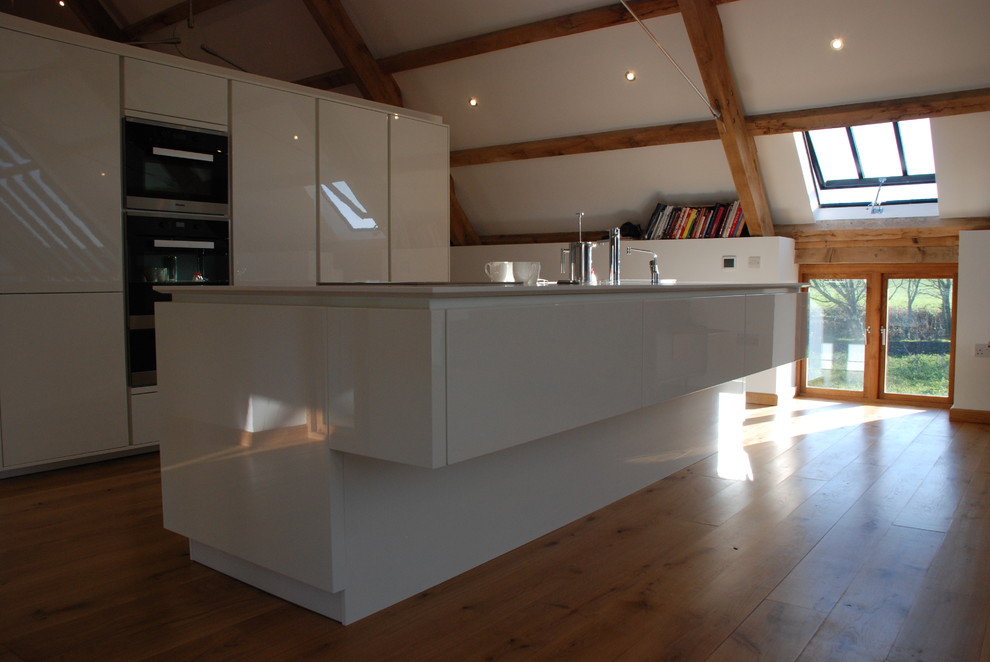 Photo of a large contemporary kitchen/diner in Cornwall with a built-in sink, flat-panel cabinets, white cabinets, composite countertops, medium hardwood flooring, an island, stainless steel appliances, a vaulted ceiling and white worktops.