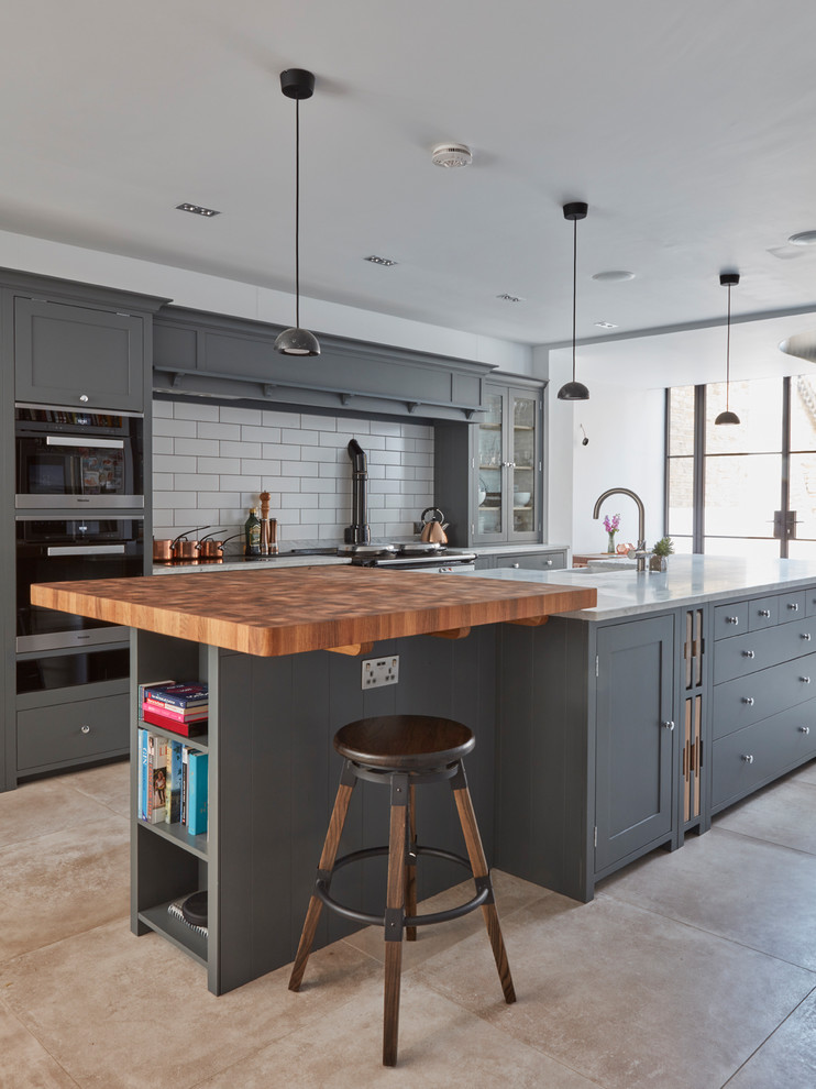 ロンドンにあるトランジショナルスタイルのおしゃれなアイランドキッチン (シェーカースタイル扉のキャビネット、グレーのキャビネット、白いキッチンパネル、サブウェイタイルのキッチンパネル、黒い調理設備、ベージュの床、白いキッチンカウンター) の写真