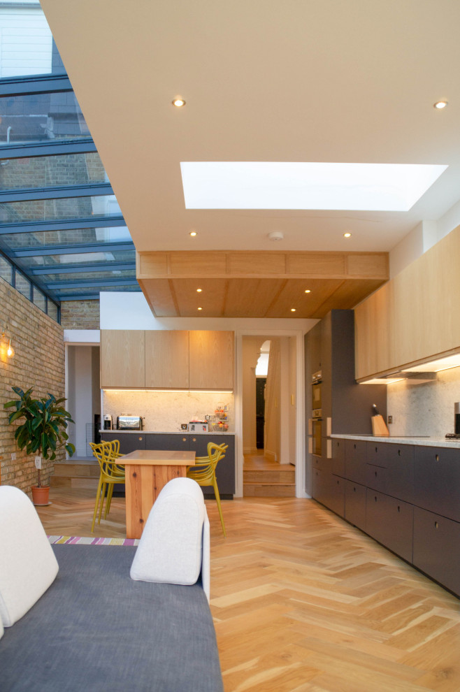 Medium sized contemporary l-shaped open plan kitchen in London with flat-panel cabinets, black cabinets, grey splashback, integrated appliances, light hardwood flooring, no island, beige floors and grey worktops.