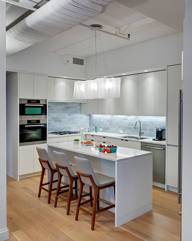 Cette image montre une cuisine américaine urbaine avec un placard à porte plane, des portes de placard blanches, une crédence bleue, un sol en bois brun, îlot, un sol marron et un plan de travail blanc.