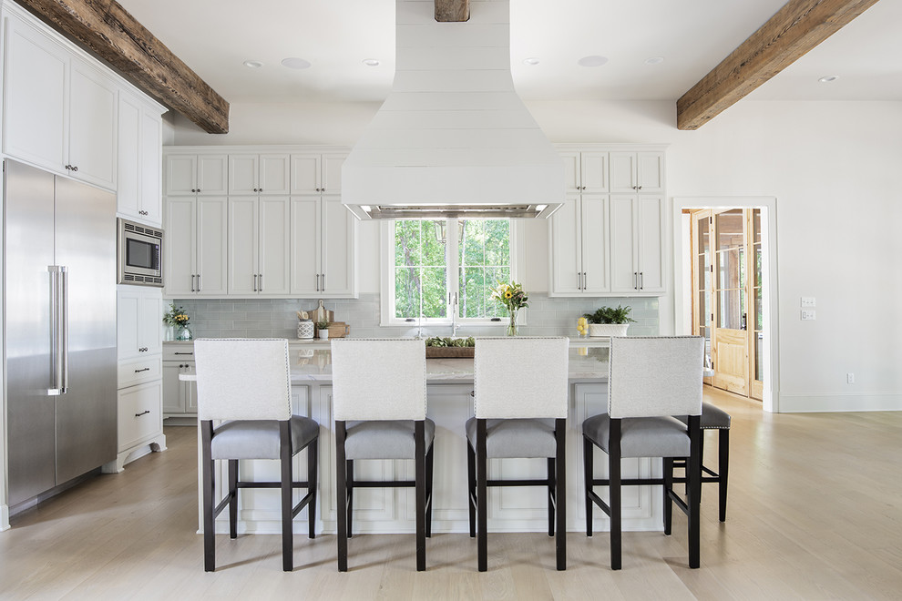 Lantlig inredning av ett vit vitt l-kök, med luckor med infälld panel, vita skåp, grått stänkskydd, rostfria vitvaror, ljust trägolv, en köksö och beiget golv