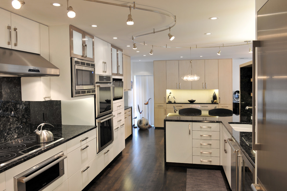Bild på ett funkis kök och matrum, med rostfria vitvaror, en undermonterad diskho, släta luckor, vita skåp, granitbänkskiva, svart stänkskydd, stänkskydd i sten, mörkt trägolv och en halv köksö