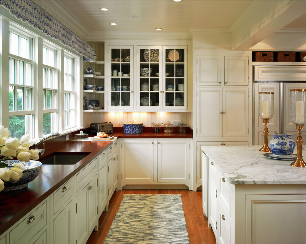 Klassisk inredning av ett kök, med en undermonterad diskho, luckor med profilerade fronter, vita skåp, träbänkskiva, integrerade vitvaror, flerfärgad stänkskydd, stänkskydd i stenkakel, mellanmörkt trägolv och en köksö