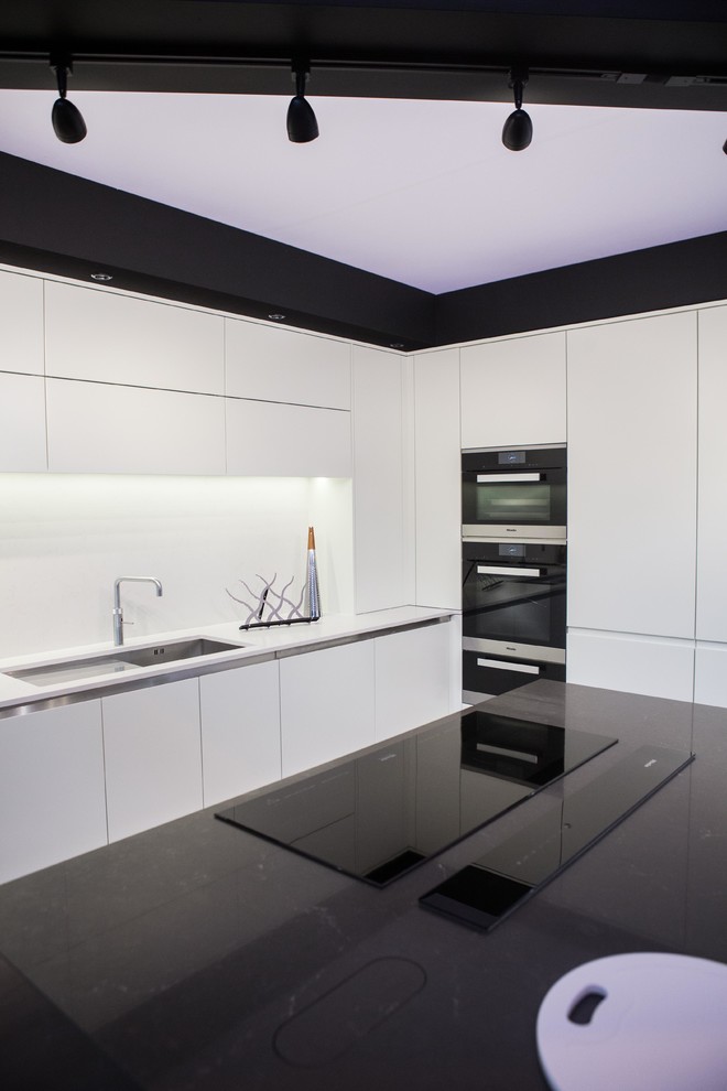 Design ideas for a large contemporary kitchen/diner in Buckinghamshire with a submerged sink, flat-panel cabinets, white cabinets, quartz worktops, white splashback, glass sheet splashback, an island and black appliances.