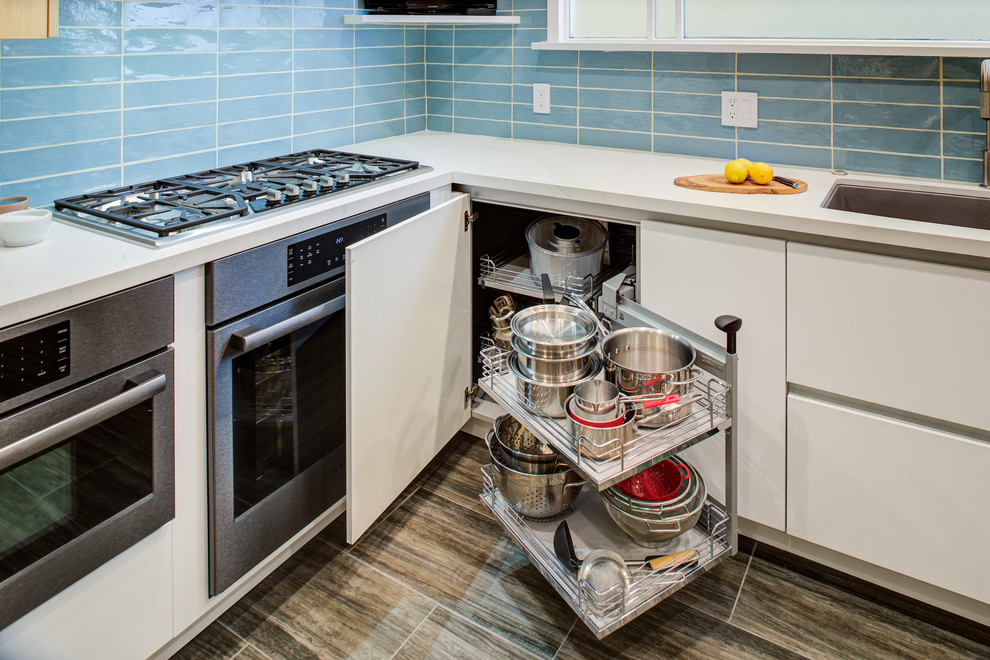 Modern inredning av ett avskilt vit vitt l-kök, med en undermonterad diskho, släta luckor, skåp i ljust trä, bänkskiva i kvarts, blått stänkskydd, stänkskydd i glaskakel, svarta vitvaror, mellanmörkt trägolv, en köksö och brunt golv