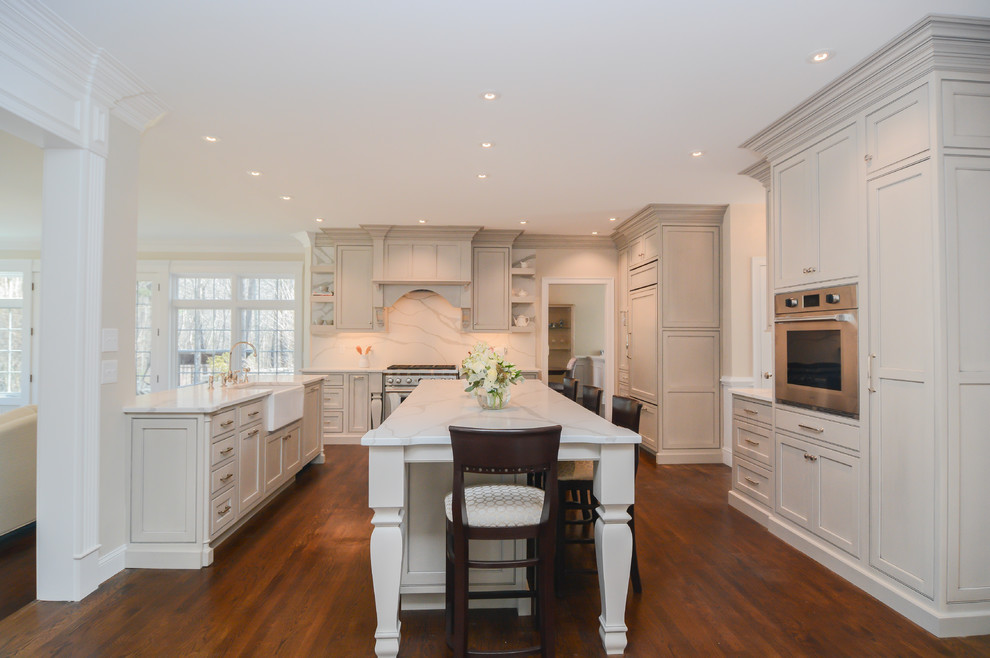 Klassisk inredning av ett mycket stort vit vitt kök med öppen planlösning, med en rustik diskho, grå skåp, bänkskiva i kvarts, vitt stänkskydd, stänkskydd i sten, rostfria vitvaror, mellanmörkt trägolv, flera köksöar, skåp i shakerstil och brunt golv