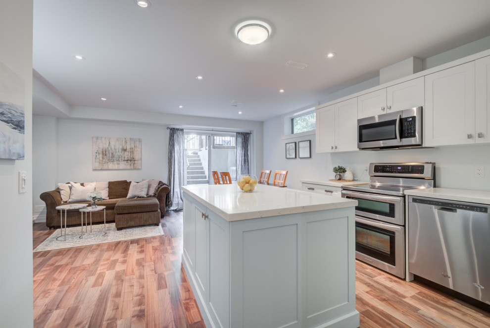 Classic open plan kitchen in Toronto with a submerged sink, shaker cabinets, white cabinets, engineered stone countertops, white splashback, stone slab splashback, stainless steel appliances, light hardwood flooring, an island, brown floors and white worktops.
