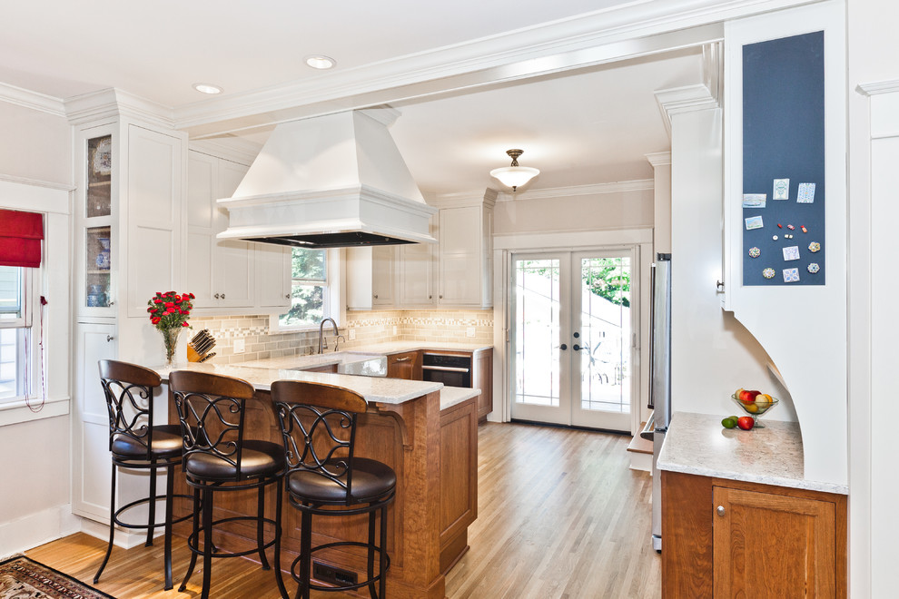 This is an example of a medium sized classic u-shaped kitchen in Portland with a belfast sink, recessed-panel cabinets, light wood cabinets, engineered stone countertops, grey splashback, ceramic splashback, stainless steel appliances, light hardwood flooring, a breakfast bar and brown floors.