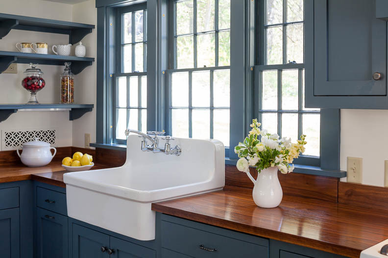 Idée de décoration pour une cuisine américaine champêtre en L avec un évier de ferme, des portes de placard bleues, un plan de travail en bois et une crédence beige.