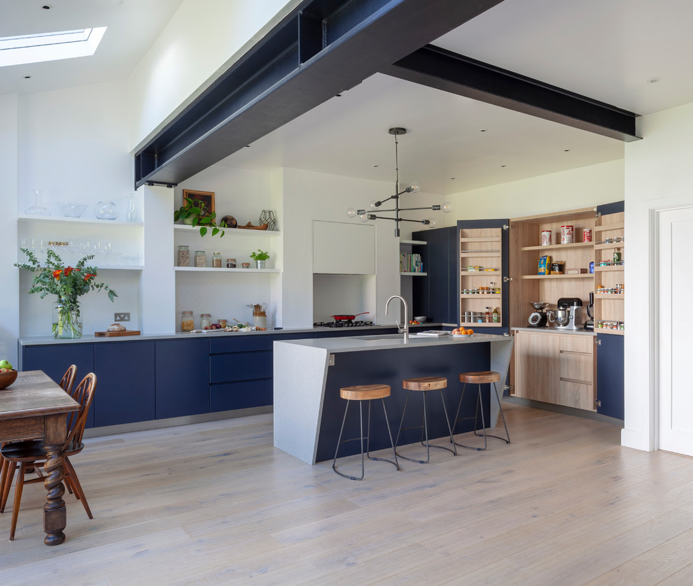 Contemporary l-shaped kitchen/diner in London with flat-panel cabinets, blue cabinets, white splashback, light hardwood flooring, an island, beige floors and grey worktops.