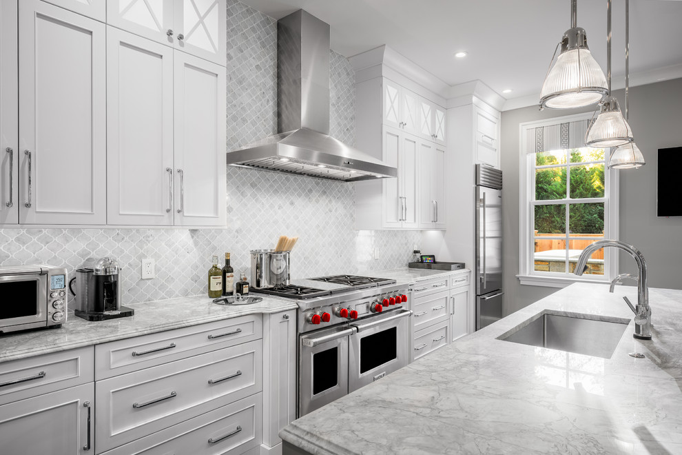 Example of a large classic galley medium tone wood floor kitchen design in Philadelphia with white cabinets, marble countertops, gray backsplash, stone tile backsplash, stainless steel appliances, an island, an undermount sink and recessed-panel cabinets