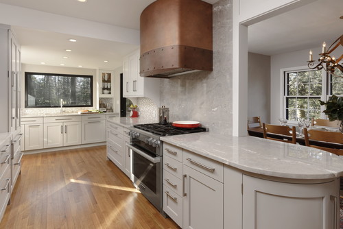 Kitchen & Bathroom Cabinets – light-gray Shaker kitchen with curved quartz peninsula, copper range hood, hardwood floor, and windowed sink.