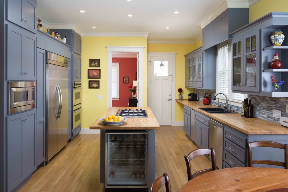 Inredning av ett modernt kök, med en dubbel diskho, luckor med glaspanel, blå skåp, träbänkskiva, blått stänkskydd, rostfria vitvaror, ljust trägolv, en köksö och stänkskydd i glaskakel