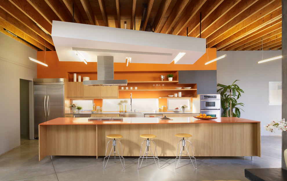 Zweizeilige Moderne Wohnküche mit Landhausspüle, flächenbündigen Schrankfronten, hellen Holzschränken, Küchenrückwand in Orange, Rückwand-Fenster, Küchengeräten aus Edelstahl, Betonboden, Kücheninsel, grauem Boden und oranger Arbeitsplatte in Los Angeles