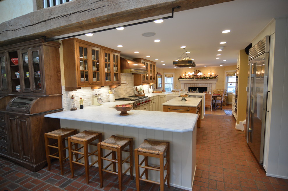 Photo of a large farmhouse u-shaped kitchen/diner in DC Metro with a belfast sink, beaded cabinets, brown cabinets, marble worktops, white splashback, ceramic splashback, stainless steel appliances, brick flooring, an island and red floors.