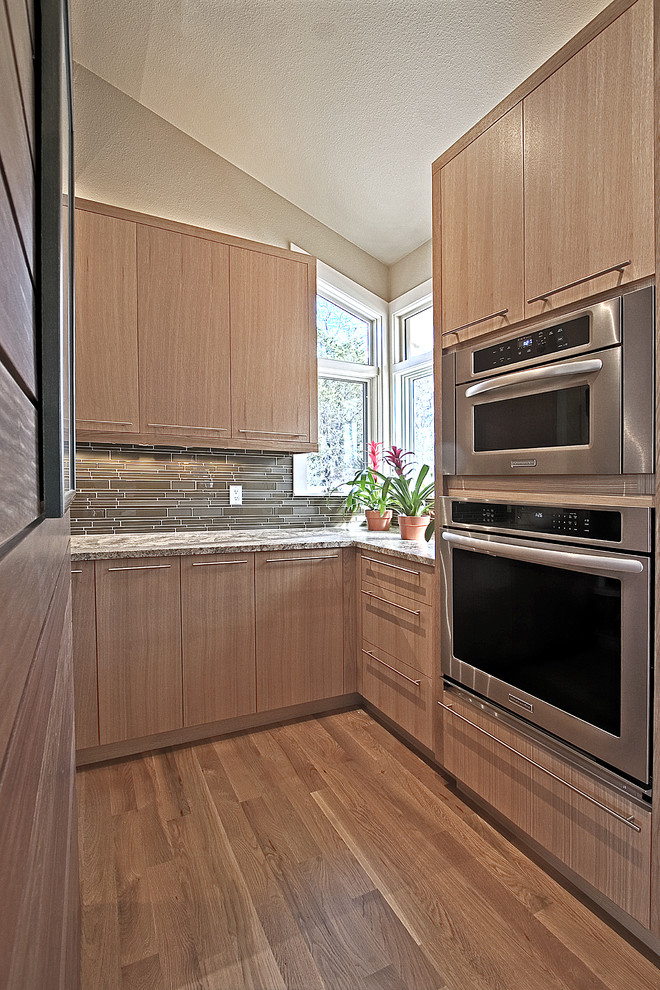 Modern inredning av ett stort kök, med en undermonterad diskho, släta luckor, bruna skåp, granitbänkskiva, grönt stänkskydd, stänkskydd i glaskakel, rostfria vitvaror, ljust trägolv, en köksö och brunt golv