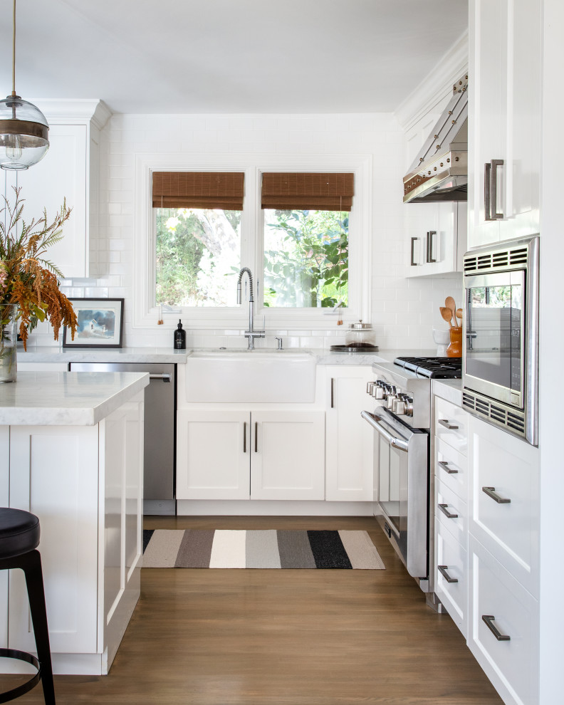Inspiration for a large traditional l-shaped kitchen in San Francisco with a belfast sink, shaker cabinets, white cabinets, white splashback, metro tiled splashback, stainless steel appliances, an island, brown floors and white worktops.