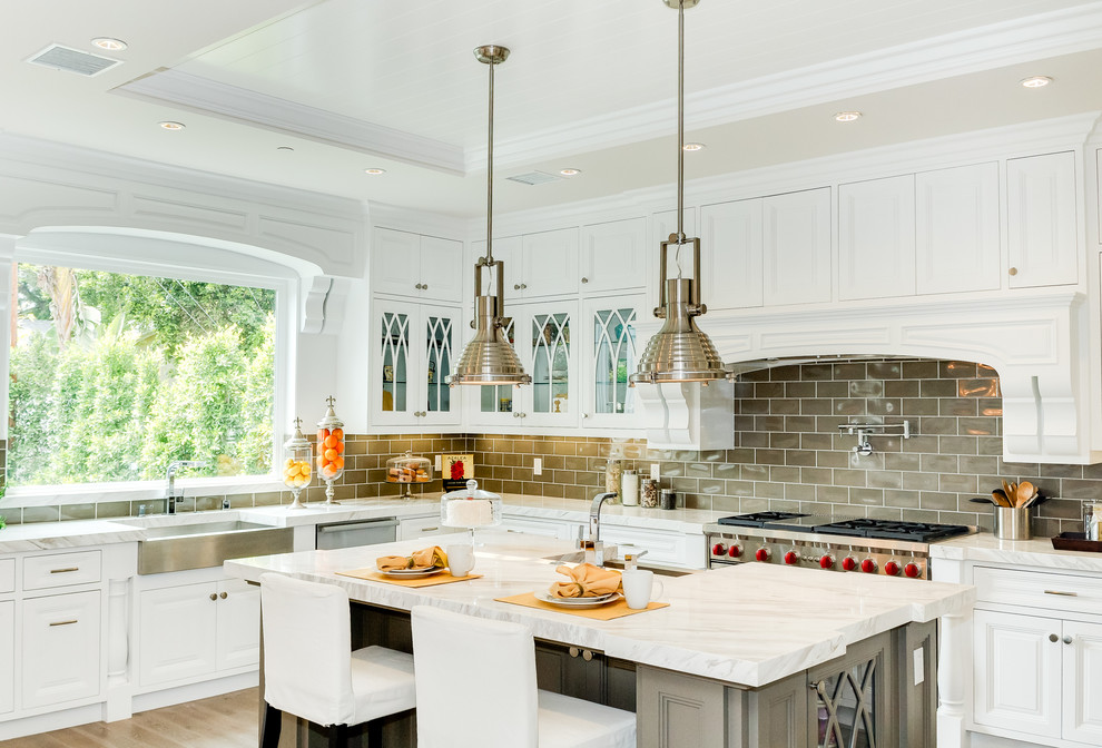 This is an example of a traditional l-shaped kitchen in Los Angeles with a belfast sink, raised-panel cabinets, white cabinets, marble worktops, grey splashback, metro tiled splashback, stainless steel appliances, light hardwood flooring and an island.
