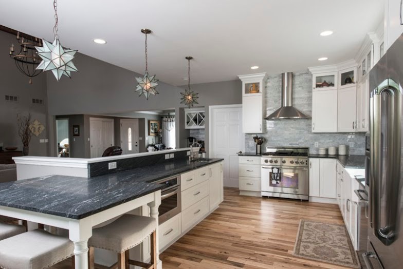 Photo of a large contemporary l-shaped open plan kitchen in Milwaukee with a belfast sink, shaker cabinets, white cabinets, engineered stone countertops, grey splashback, marble splashback, stainless steel appliances, light hardwood flooring and an island.