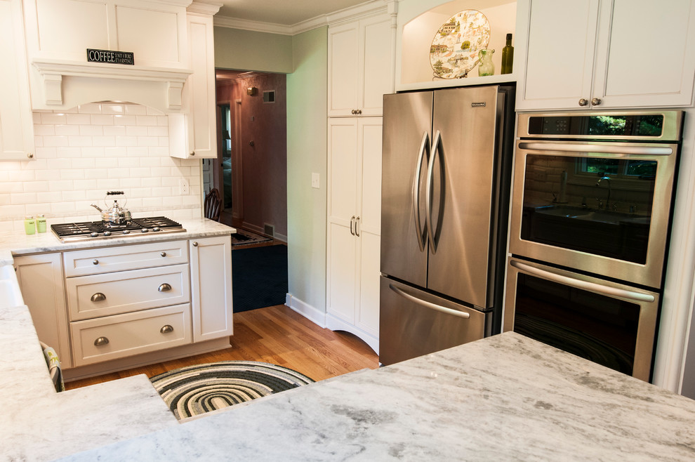 Inspiration for a small transitional u-shaped medium tone wood floor enclosed kitchen remodel in Milwaukee with a farmhouse sink, shaker cabinets, white cabinets, granite countertops, white backsplash, subway tile backsplash, stainless steel appliances and a peninsula