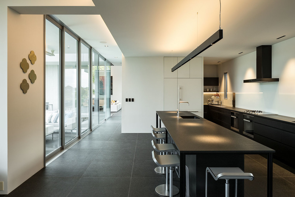 This is an example of a modern cream and black l-shaped kitchen/diner in Auckland with black cabinets, white splashback, glass sheet splashback, an island, black floors, black worktops, ceramic flooring, a submerged sink, flat-panel cabinets and integrated appliances.