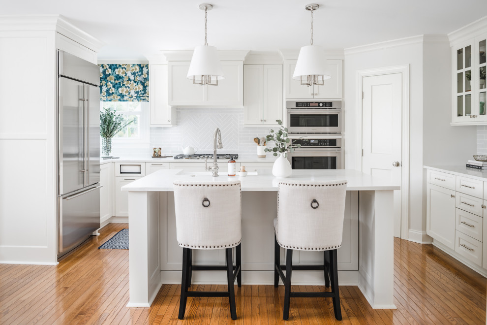 This is an example of a medium sized traditional u-shaped kitchen in Charlotte with a belfast sink, shaker cabinets, white cabinets, engineered stone countertops, grey splashback, porcelain splashback, stainless steel appliances, medium hardwood flooring, an island, brown floors and white worktops.