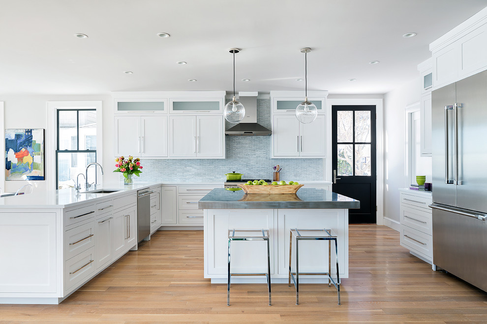 This is an example of a coastal l-shaped kitchen in New York with a submerged sink, shaker cabinets, white cabinets, grey splashback, stainless steel appliances, light hardwood flooring and an island.