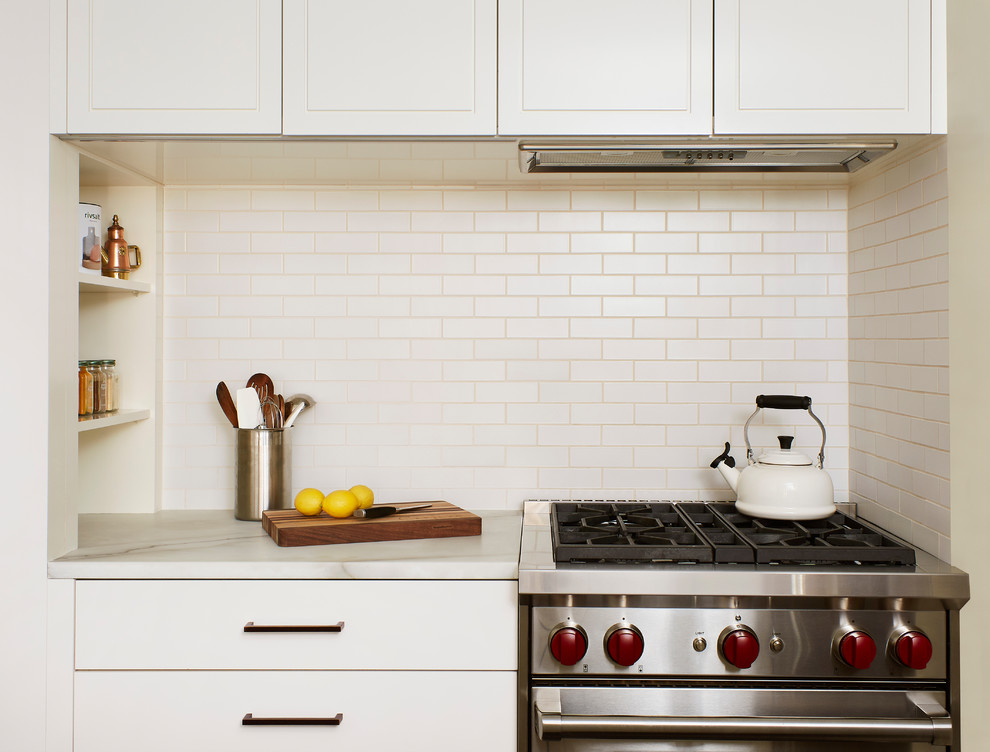 Photo of a small contemporary u-shaped kitchen/diner in New York with flat-panel cabinets, white splashback, ceramic splashback, stainless steel appliances, an island and white worktops.
