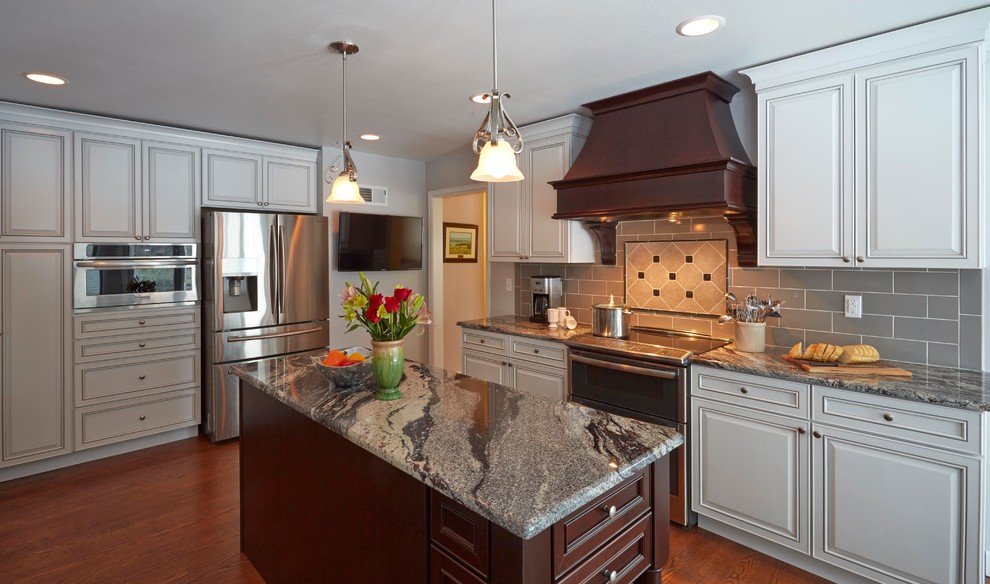Klassisk inredning av ett mellanstort kök, med en undermonterad diskho, luckor med upphöjd panel, vita skåp, granitbänkskiva, grått stänkskydd, stänkskydd i cementkakel, rostfria vitvaror, mellanmörkt trägolv och flera köksöar