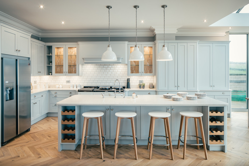 ハートフォードシャーにある広いトランジショナルスタイルのおしゃれなキッチン (青いキャビネット、シェーカースタイル扉のキャビネット、白いキッチンパネル、サブウェイタイルのキッチンパネル、シルバーの調理設備、淡色無垢フローリング、ベージュの床) の写真