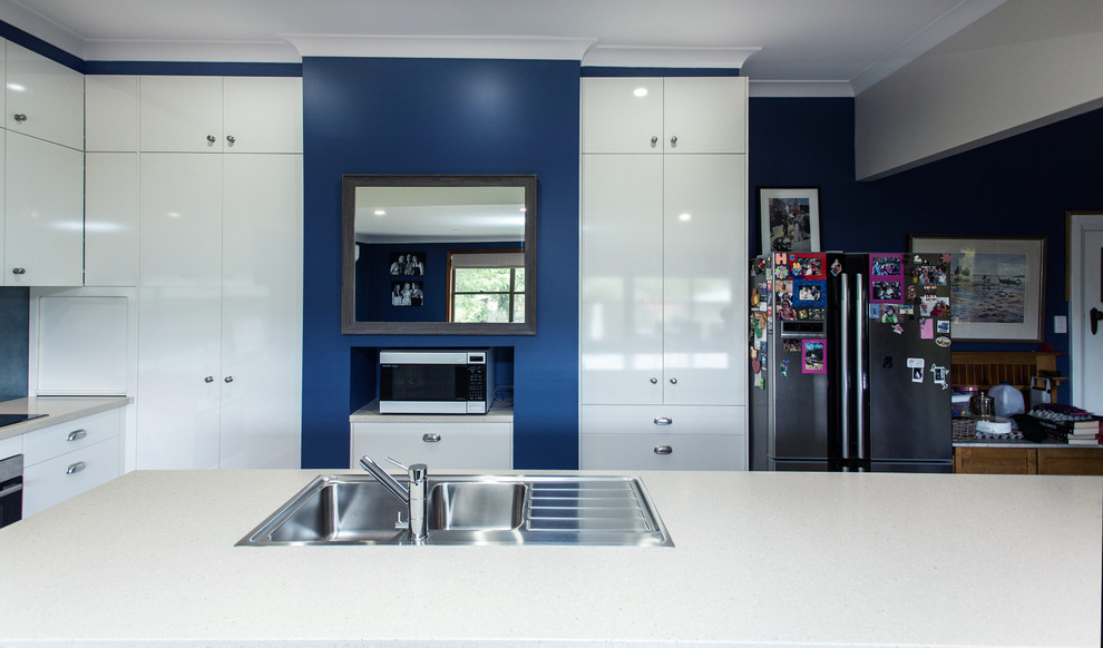 This is an example of a small contemporary galley open plan kitchen in Other with a built-in sink, flat-panel cabinets, white cabinets, composite countertops, blue splashback, glass sheet splashback, stainless steel appliances, medium hardwood flooring, a breakfast bar, brown floors and white worktops.