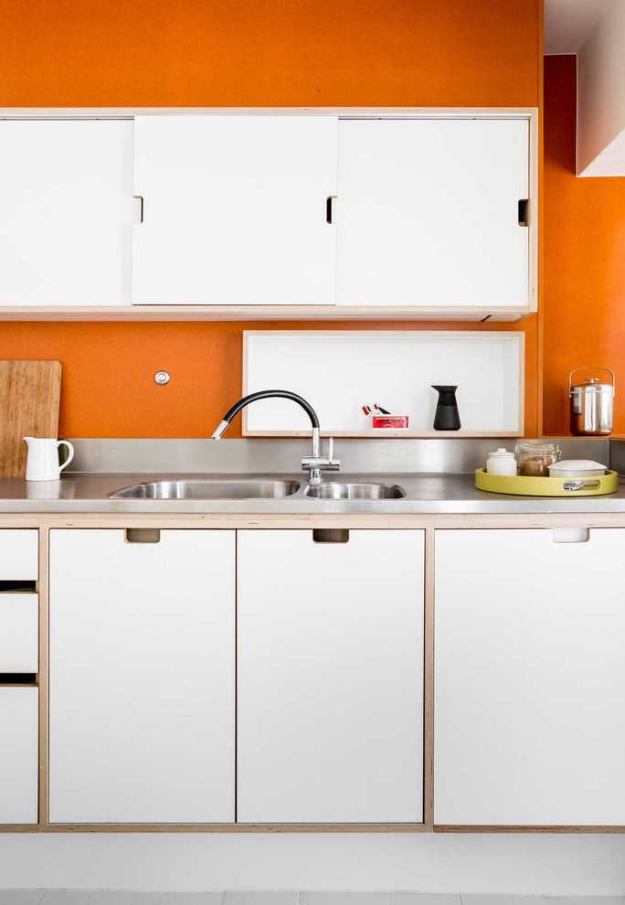 Inspiration for a medium sized contemporary u-shaped enclosed kitchen in Other with flat-panel cabinets, white cabinets, stainless steel worktops, stainless steel appliances, light hardwood flooring, an island, white floors, a built-in sink and orange splashback.