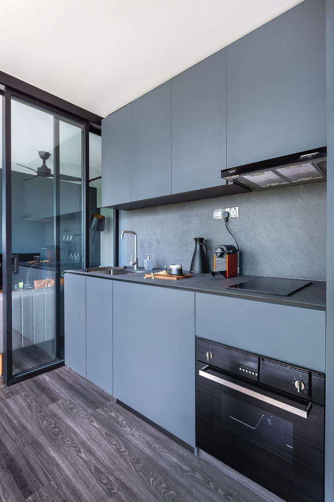 Inredning av ett modernt grå linjärt grått kök, med en enkel diskho, släta luckor, blå skåp, blått stänkskydd, svarta vitvaror, mörkt trägolv och brunt golv