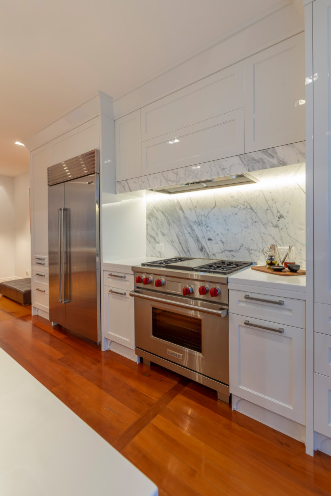 Example of a mid-sized transitional single-wall medium tone wood floor and brown floor eat-in kitchen design in Auckland with an undermount sink, recessed-panel cabinets, white cabinets, solid surface countertops, gray backsplash, marble backsplash, stainless steel appliances, an island and white countertops